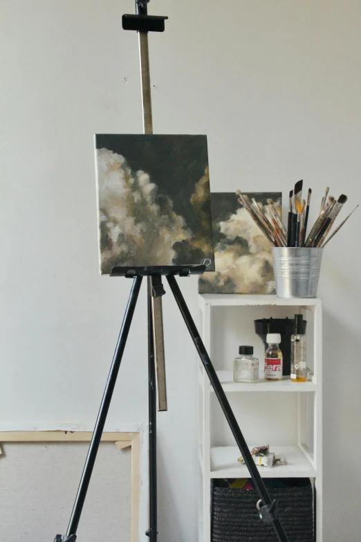 a painting sitting on top of a tripod next to a shelf, inspired by Emil Carlsen, unsplash, academic art, tall fluffy clouds, charcoal and champagne, studio background, in a studio