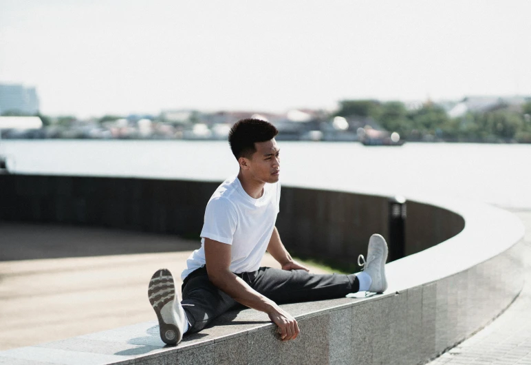 a man sitting on a wall next to a body of water, happening, workout, set on singaporean aesthetic, portrait image, plain stretching into distance