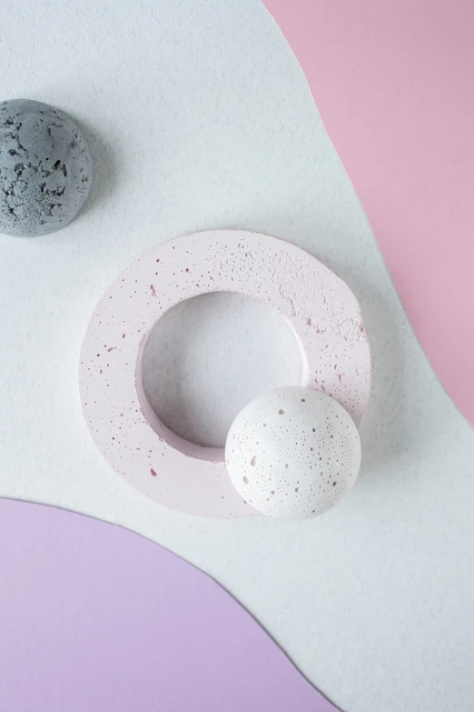 a white object sitting on top of a pink and purple surface, inspired by jeonseok lee, trending on unsplash, concrete art, shaped like torus ring, mini planets, detailed letters, black white pastel pink
