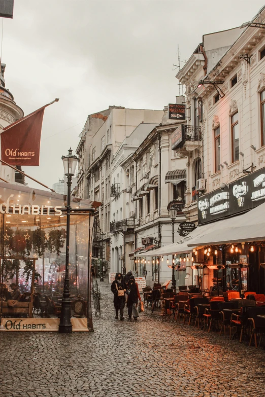 a couple of people walking down a cobblestone street, pexels contest winner, art nouveau, rostov city, people sitting at tables, warm street lights store front, rainy afternoon