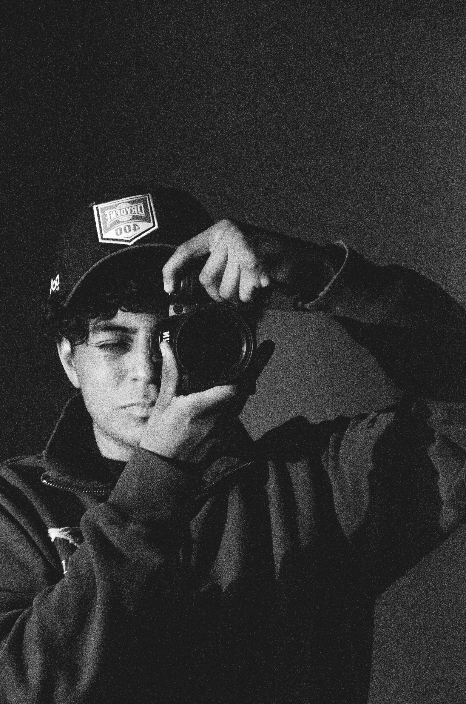a black and white photo of a man holding a camera, a black and white photo, inspired by Ion Andreescu, wearing a backwards baseball cap, young prince, highly reflective, ariel perez