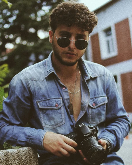 a man sitting on a wall holding a camera, an album cover, trending on pexels, jewish young man with glasses, wearing double denim, headshot profile picture, ahmad merheb