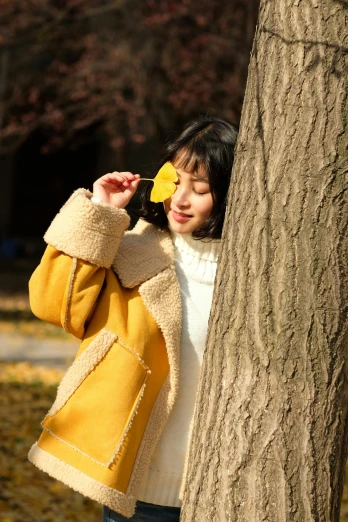 a woman leaning against a tree with a flower in her hand, inspired by Ye Xin, trending on pexels, yellow fur, sunny winter day, 🍁 cute, warm color clothes