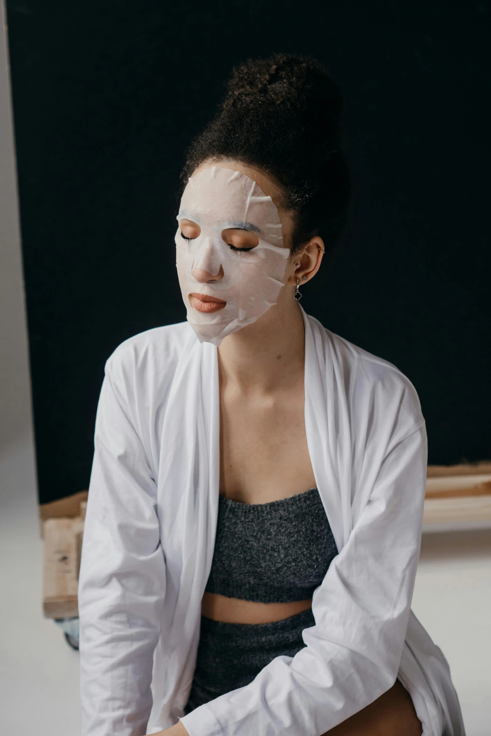 a woman sitting on a stool with a face mask on, inspired by Marina Abramović, trending on pexels, renaissance, skincare, half body portrait, wearing a grey robe, square facial structure