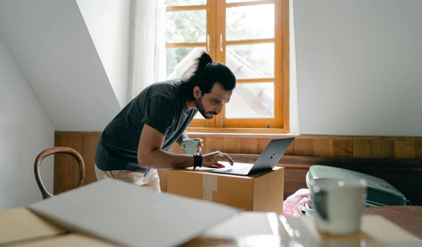 a man sitting at a table working on a laptop, pexels contest winner, hurufiyya, delivering parsel box, standing near a window, inspect in inventory image, sitting on edge of bed