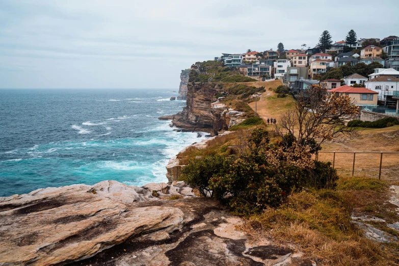 a group of houses sitting on top of a cliff next to the ocean, inspired by Sydney Carline, pexels contest winner, manly, fall season, kodak photo, people walking around