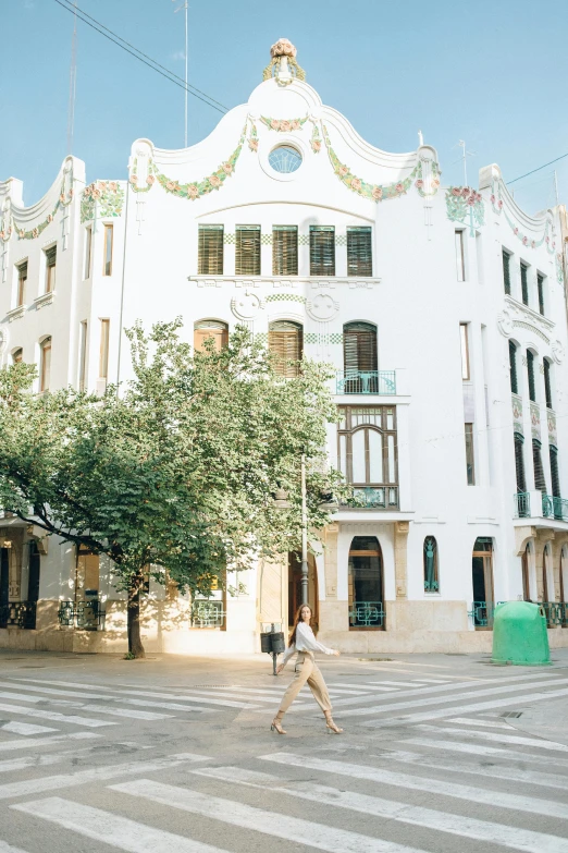 a woman walking across a crosswalk in front of a building, inspired by Tomàs Barceló, unsplash, art nouveau, wide view, 2 5 6 x 2 5 6 pixels, white houses, monserrat gudiol