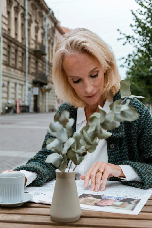 a woman sitting at a table reading a newspaper, a photo, by Grytė Pintukaitė, pexels contest winner, with soft bushes, wearing a green sweater, in the city, a blond