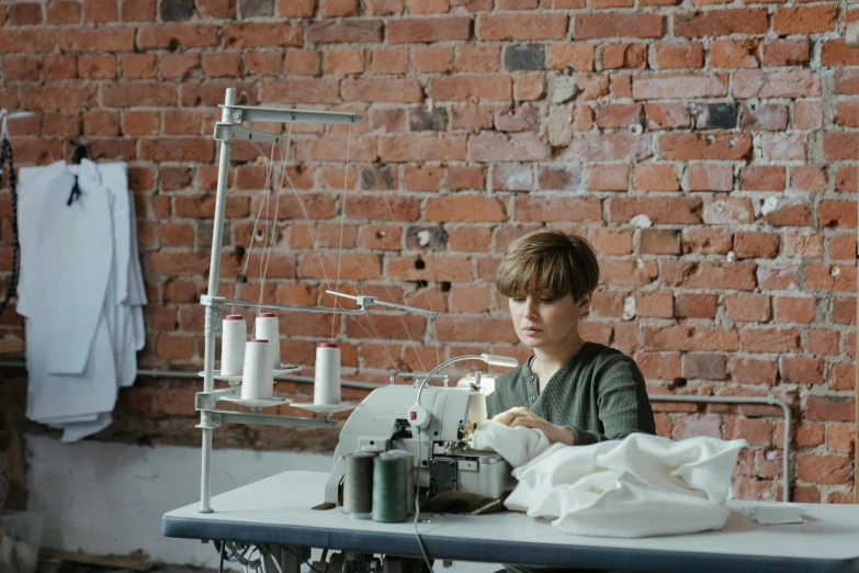 a woman sitting at a sewing machine in front of a brick wall, work clothes, on a white table, bulky build, ignant