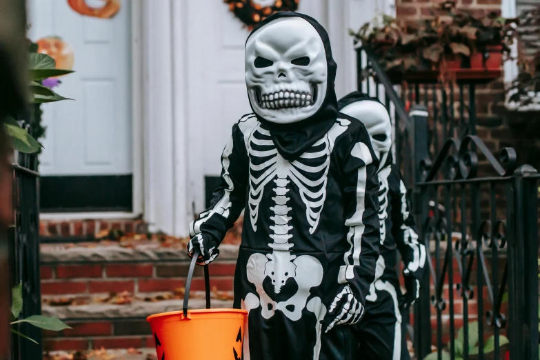 a person in a skeleton costume holding a bucket, by Julia Pishtar, pexels, children's, 💋 💄 👠 👗, in front of the house, boys