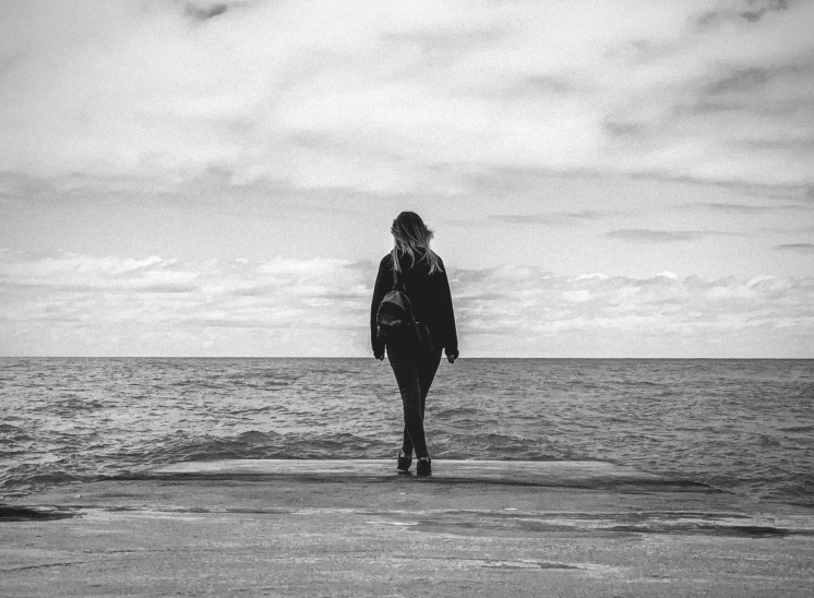 a woman standing on top of a beach next to the ocean, a black and white photo, unsplash, minimalism, in a black hoodie, feeling miserable, walking down, alternate album cover