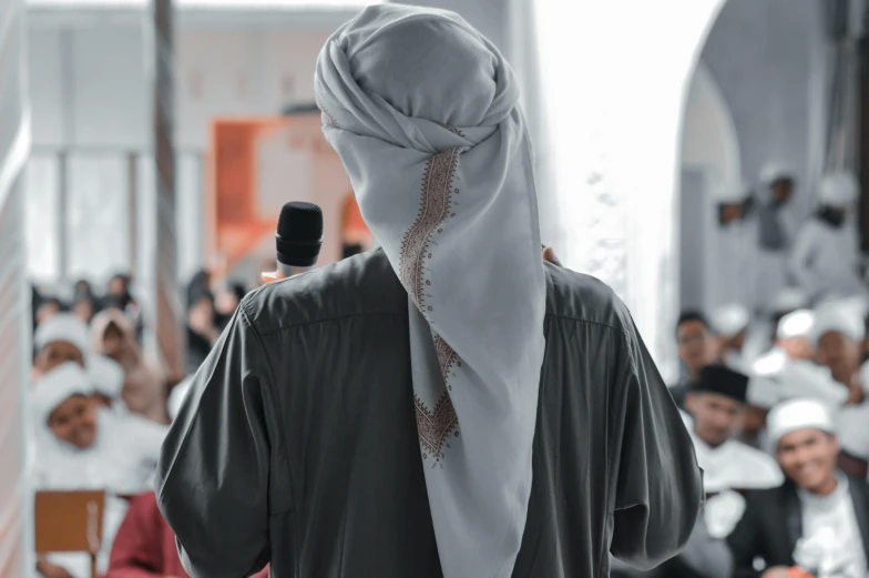 a man standing in front of a crowd of people, inspired by Abdullah Gërguri, pexels contest winner, hurufiyya, wearing white silk hood, standing in class, view from back, holding microphone