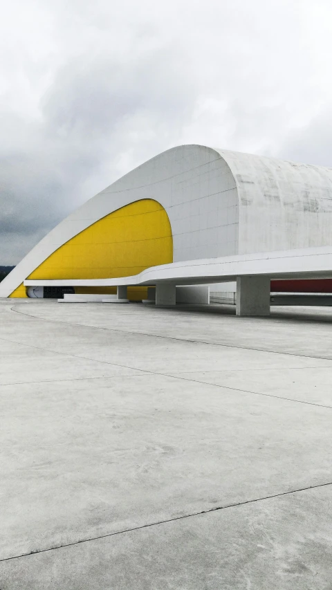 a yellow and white building sitting on top of a cement ground, oscar niemeyer, spaceship hangar, on a gray background, colorful caparisons