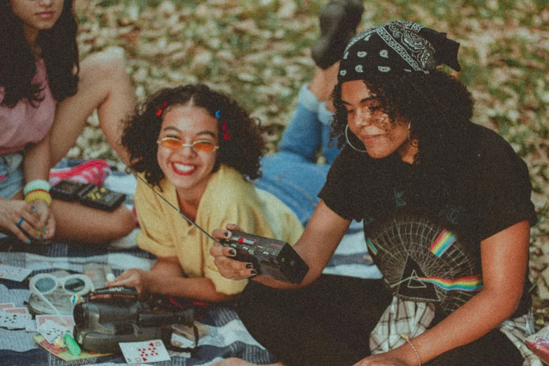 a group of women sitting on top of a blanket, a polaroid photo, trending on pexels, afrofuturism, twins playing video games, having a picnic, promotional image, controllers
