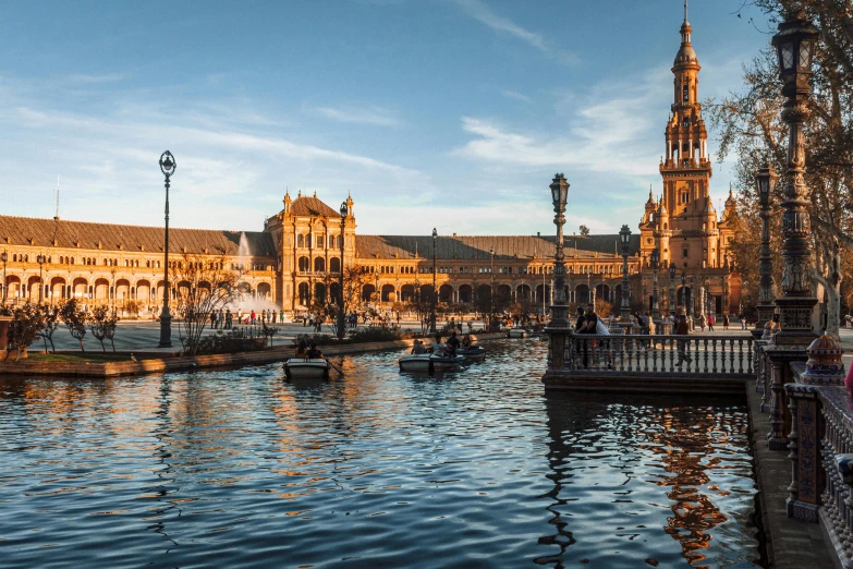 a group of people sitting on a bench next to a body of water, pexels contest winner, baroque, seville, dry archways and spires, docks, warm glow