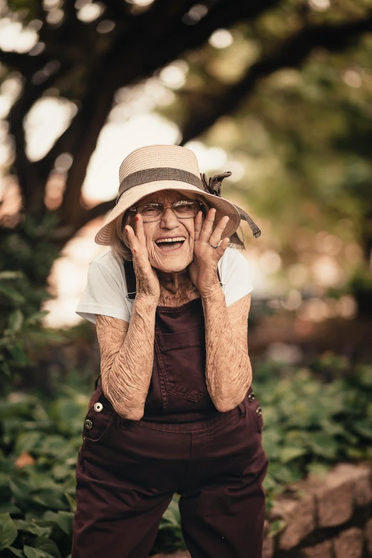 an older woman holding her hands up to her face, by Joze Ciuha, pexels contest winner, wearing a straw hat and overalls, in the garden, tattooed, goofy smile