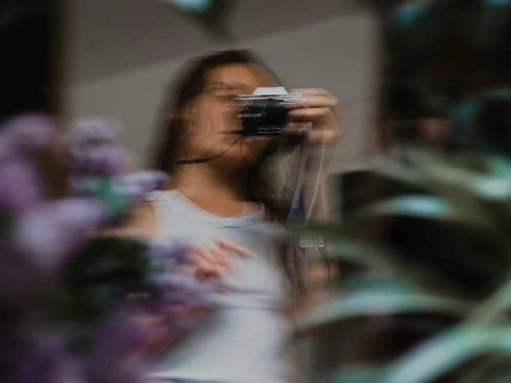 a woman taking a picture with a camera, by Emma Andijewska, visual art, blurry footage, flowers around, reflecting, looking outside