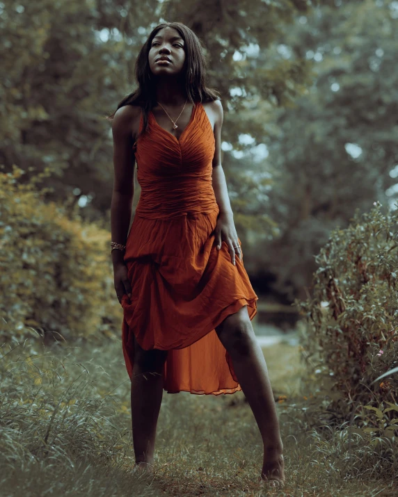 a woman in an orange dress standing in the woods, an album cover, unsplash, dark brown skin, about to step on you, low quality photo, aggressive pose