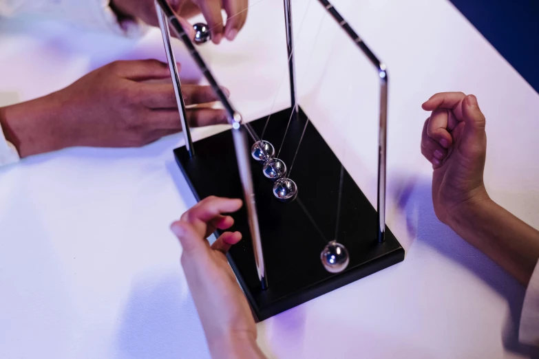 a group of people playing with pendulums on a table, inspired by Matteo Pérez, unsplash, kinetic art, ultra realistic, black 3 d cuboid device, bridge between the worlds, plating
