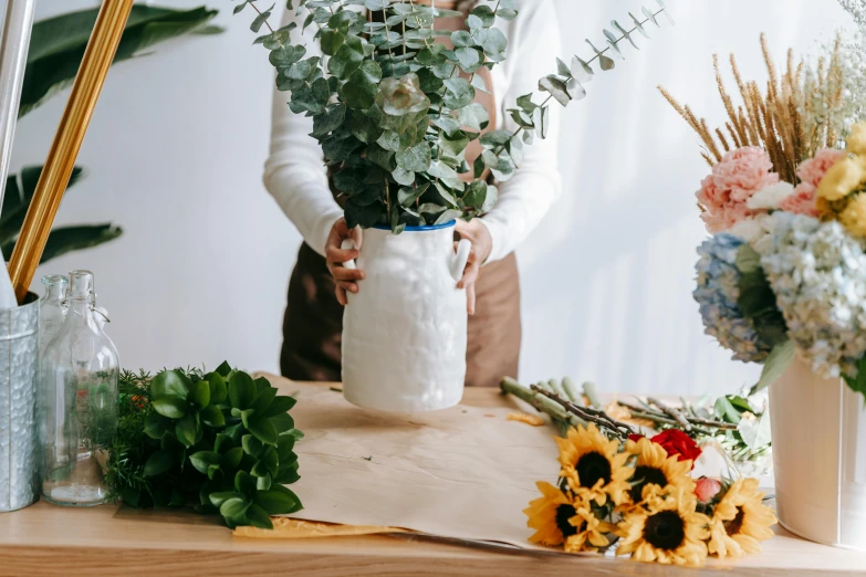 a woman arranging flowers in a vase on a table, trending on pexels, giant flowers, white vase, professional product photo, with a bunch of stuff