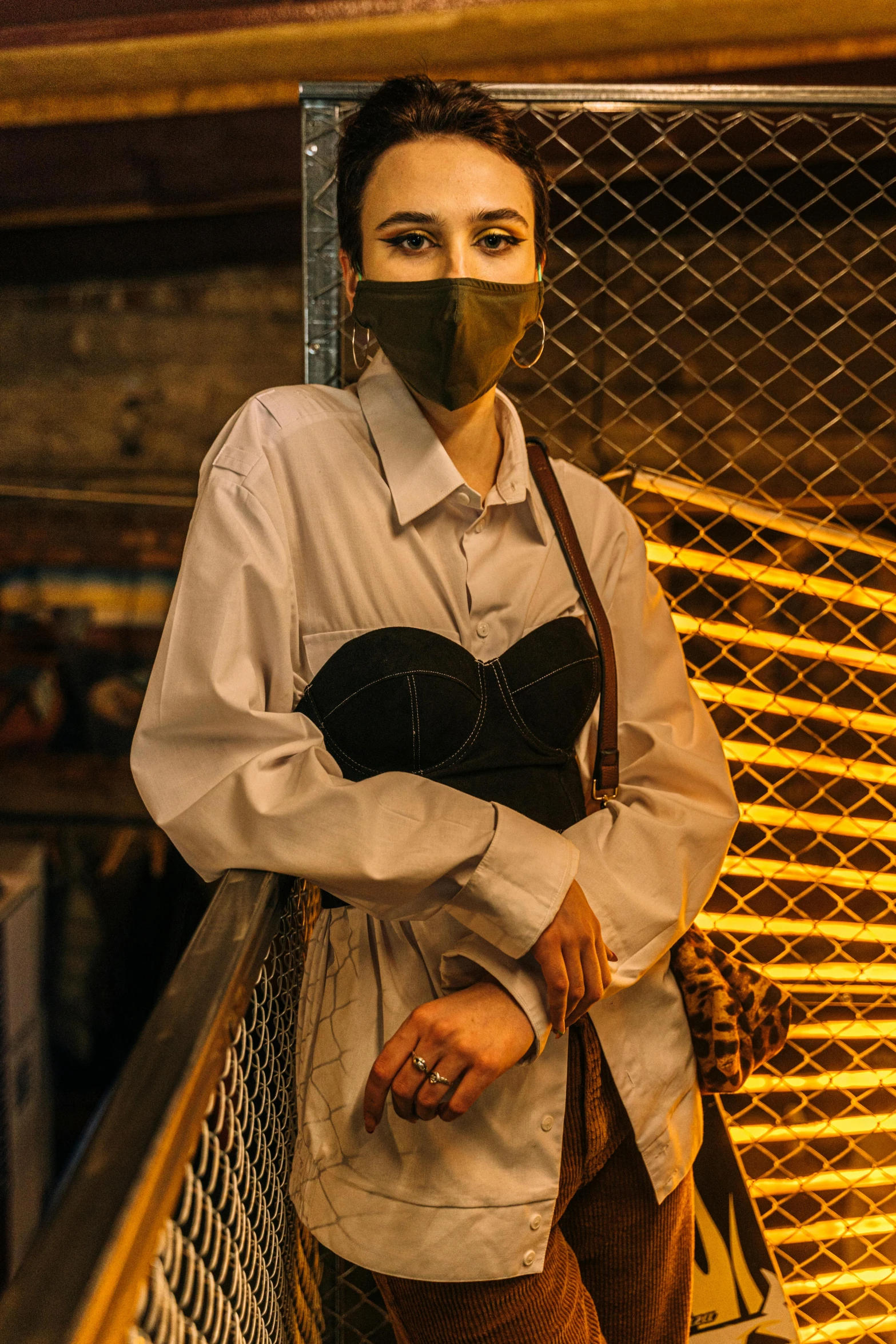 a woman wearing a face mask standing in front of a fence, inspired by Elsa Bleda, renaissance, very smoky cyberpunk paris bar, in style of lam manh, wearing a light shirt, warm moody lighting