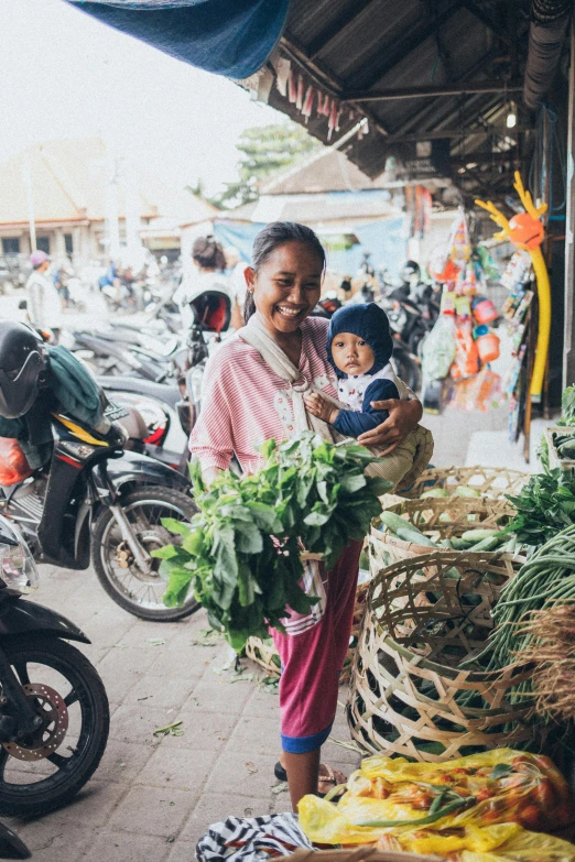 a woman holding a baby standing in front of a market, greenery, riding a motorbike, bali, square