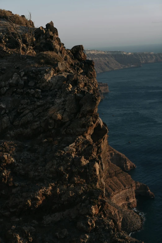 a man standing on top of a cliff next to the ocean, by Alexis Grimou, santorini, low quality photo, late summer evening, brown