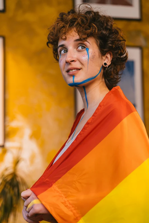 a woman holding a rainbow flag in front of a yellow wall, by Julia Pishtar, antipodeans, face painting, blue and orange, with a hurt expression, a handsome