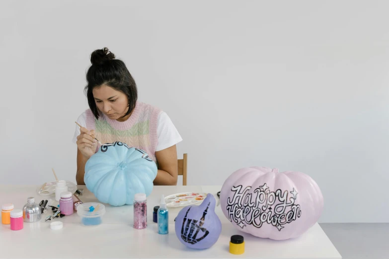 a woman sitting at a table painting a pumpkin, by Olivia Peguero, flowing lettering, blue and purple scheme, bubblegum body, hand painted textures on model