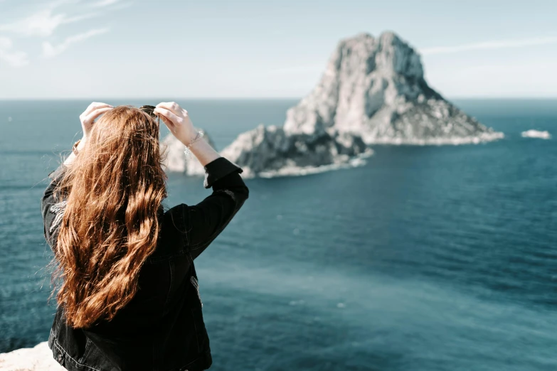 a woman standing on top of a cliff next to the ocean, by Kristian Zahrtmann, pexels contest winner, ibiza, her hair blowing in the wind, thumbnail, looking outside