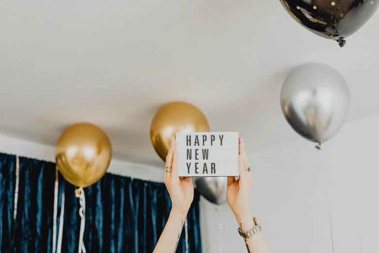 a woman holding up a sign that says happy new year, a polaroid photo, by Julia Pishtar, trending on pexels, happening, party balloons, background image, white + blue + gold + black, decoration