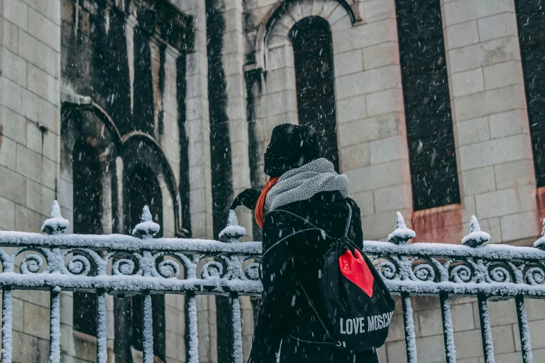 a person standing in front of a building in the snow, black and white with red hearts, a backpack, background image, lovecratian