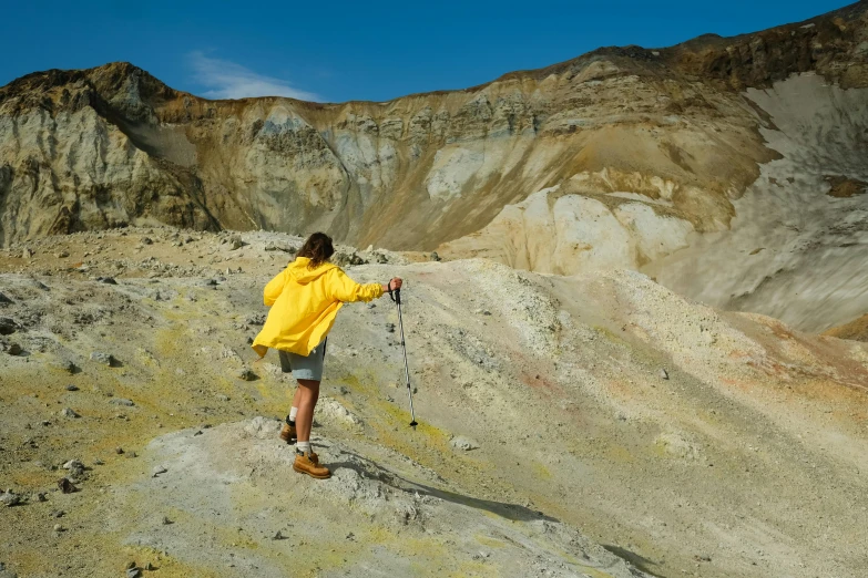 a man in a yellow jacket standing on top of a mountain, by Hallsteinn Sigurðsson, unsplash, figuration libre, bisti badlands, with a walking cane, pastels, white shorts and hiking boots