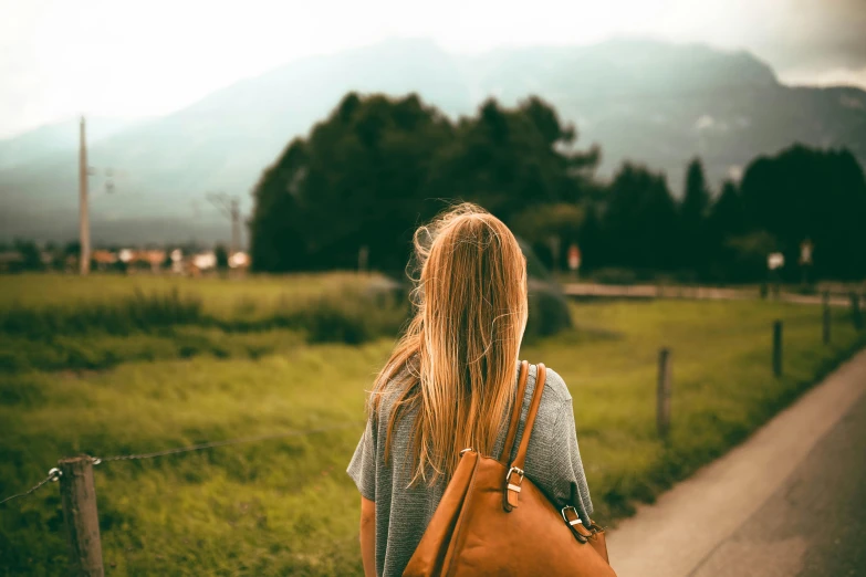 a woman walking down a road carrying a brown bag, a picture, trending on pexels, meadow in the background, messy blond hair, distant mountains, background image