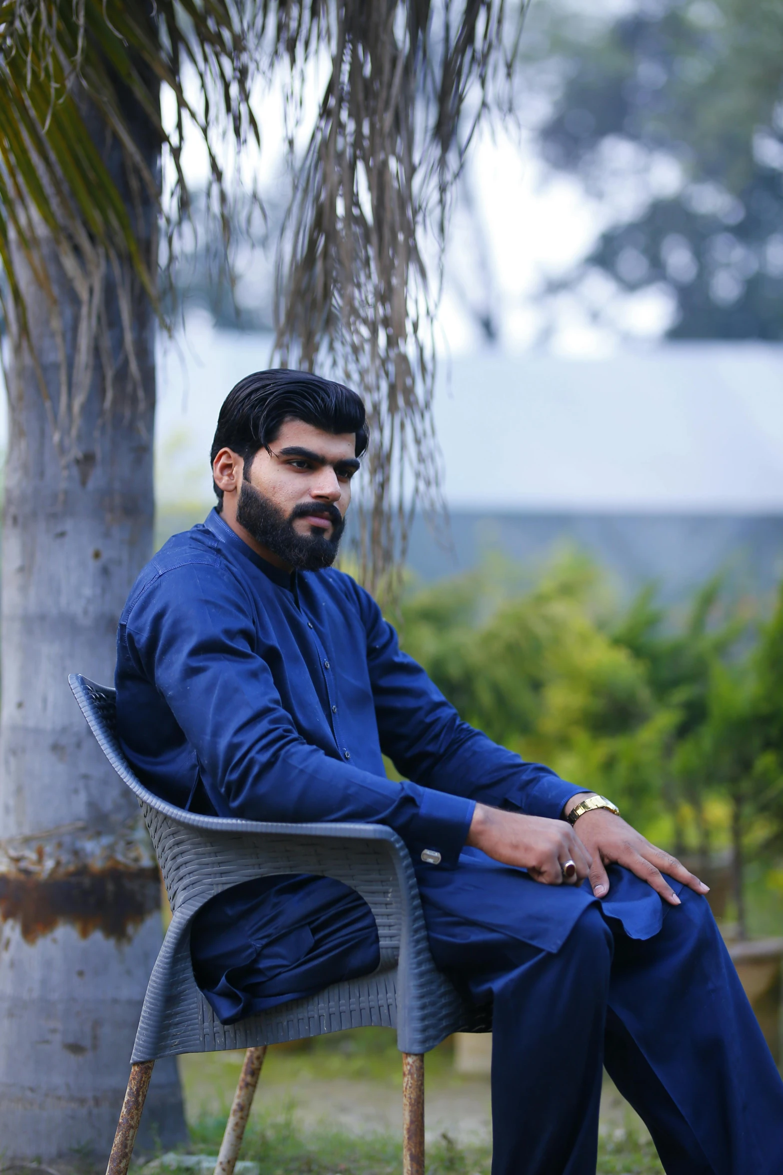 a man sitting in a chair next to a palm tree, by Riza Abbasi, pexels contest winner, hurufiyya, very attractive man with beard, in a navy blue sweater, desi, trimmed