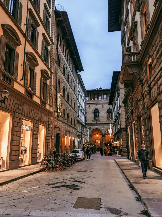 a couple of people walking down a street next to tall buildings, renaissance, florence, warm street lights store front, profile image