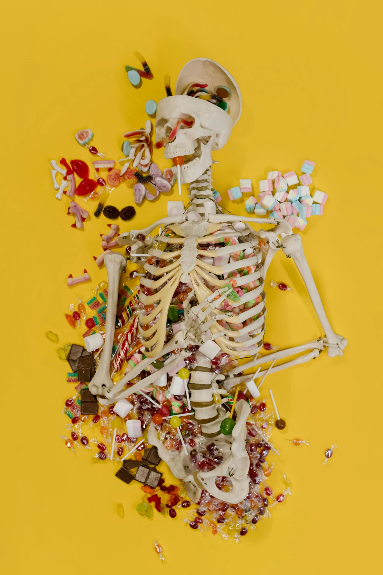 a skeleton sitting on top of a pile of candy, an album cover, by Damien Hirst, pexels, conceptual art, yellow, healthcare, rib cage, lollipop