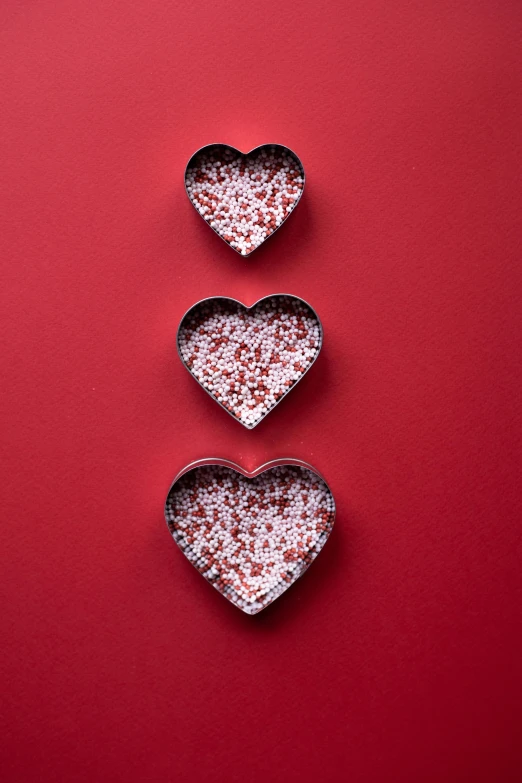 three heart shaped chocolates on a red background, by Lucette Barker, pexels, 3d flat layered paper shadow box, speckled, made out of shiny white metal, can