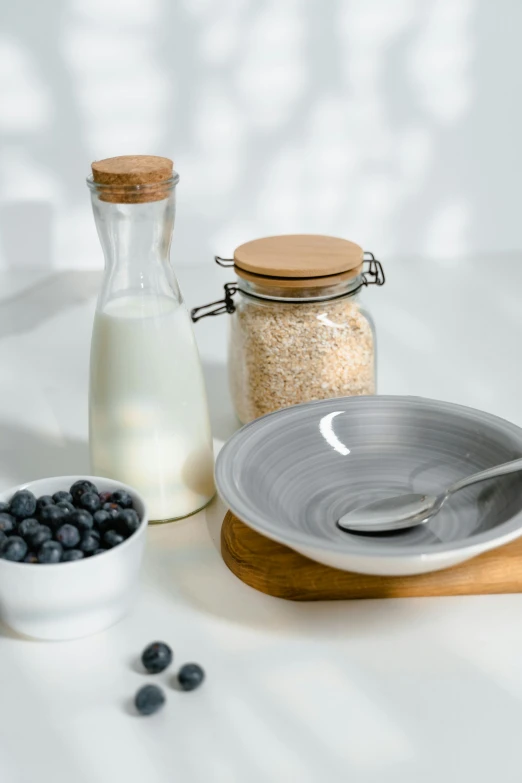 a table topped with a bowl of blueberries next to a bottle of milk, unsplash, cereal, on grey background, jar on a shelf, on a wooden plate