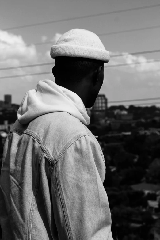 a black and white photo of a man looking out over a city, by Afewerk Tekle, hat and hoodie, an all white human, rear-shot, african man