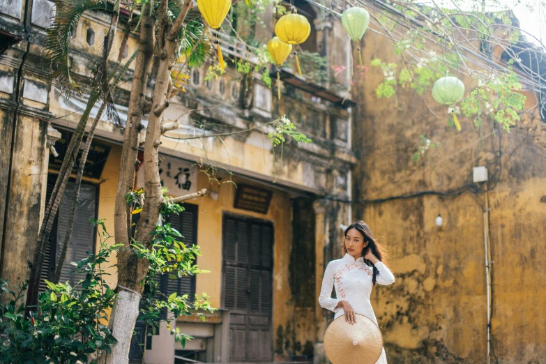 a woman standing in front of a yellow building, inspired by Ruth Jên, happening, with paper lanterns, vietnamese woman, background image, shady alleys