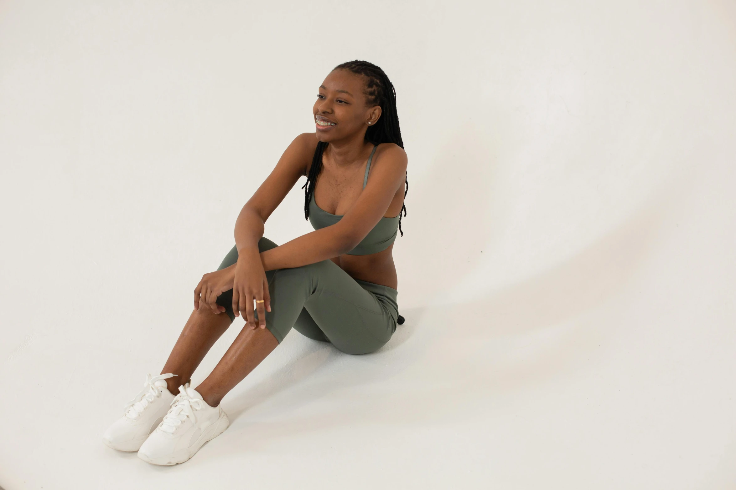 a woman sitting on the ground with her legs crossed, trending on pexels, figuration libre, olive green, sports bra, ( ( dark skin ) ), in front of white back drop