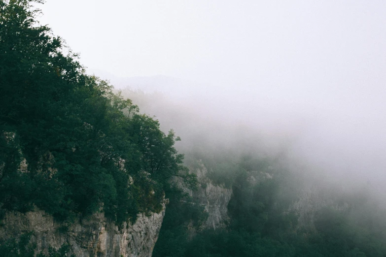 a boat sitting on top of a river next to a cliff, inspired by Elsa Bleda, pexels contest winner, romanticism, foggy heavy rain, over the tree tops, view from below, light grey mist