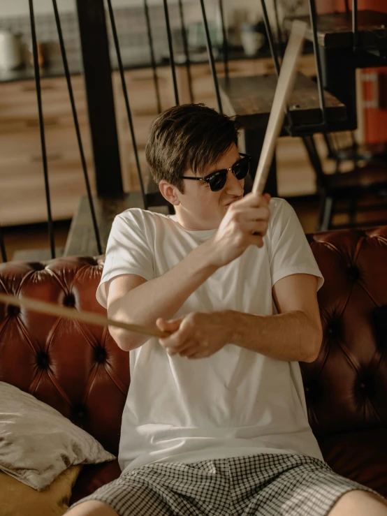 a man sitting on a couch playing drums, by Robbie Trevino, pexels contest winner, photorealism, with sunglass, dressed in a white t shirt, declan mckenna, holding a whip