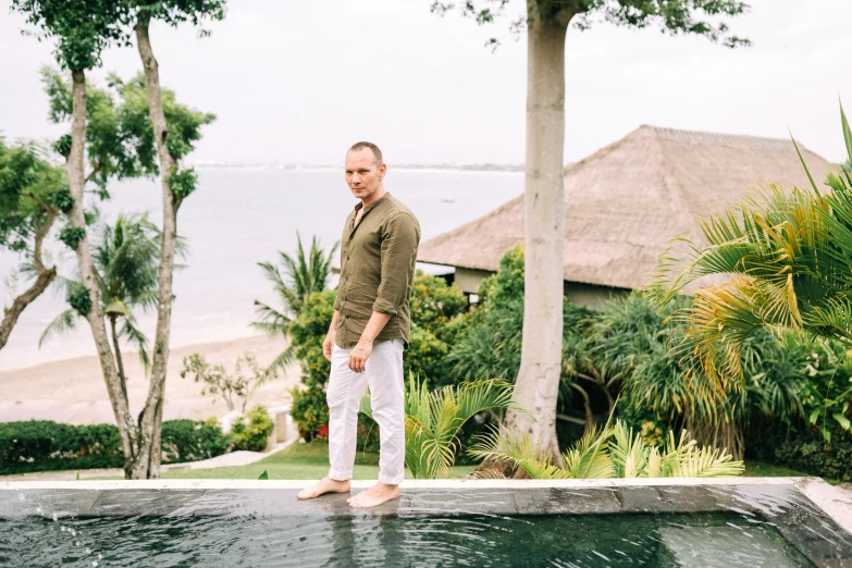 a man standing on the edge of a pool, a portrait, happening, wearing a linen shirt, bali, jony ive, tsunami behind him