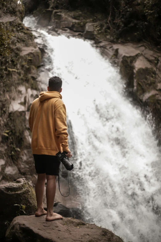 a man standing on top of a rock next to a waterfall, taking a picture, casually dressed, drenched clothing, liquid gold