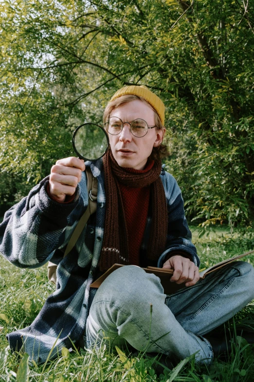 a man sitting in the grass holding a magnifying glass, an album cover, by Simon Marmion, round glasses potter, in woods, where's wally, wes anderson color palette