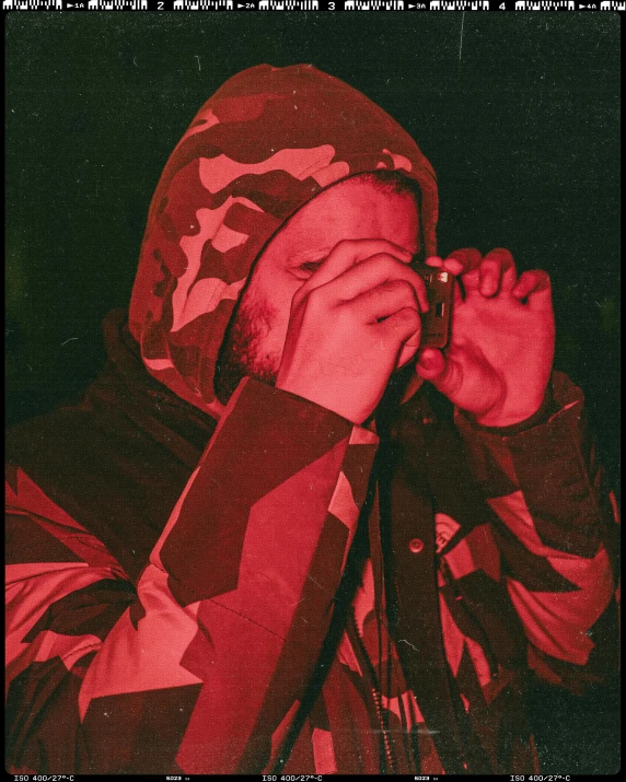 a man in a hoodie talking on a cell phone, an album cover, by Attila Meszlenyi, red tint, wearing camo, one eye red, 3 5 mm pointing up