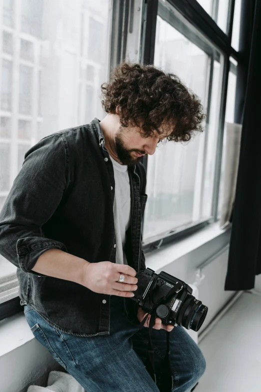 a man sitting on a window sill holding a camera, a picture, by Daniel Seghers, halfbody headshot, scruffy man, looking down on the camera, holding a dslr camera