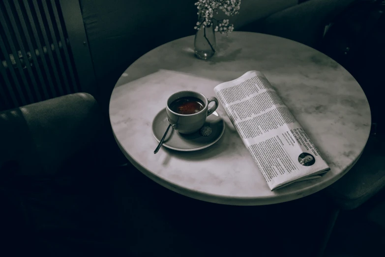 a newspaper sitting on top of a table next to a cup of coffee, inspired by Brassaï, pexels contest winner, private press, chilly dark mood, marble table, well - dressed, cafe tables
